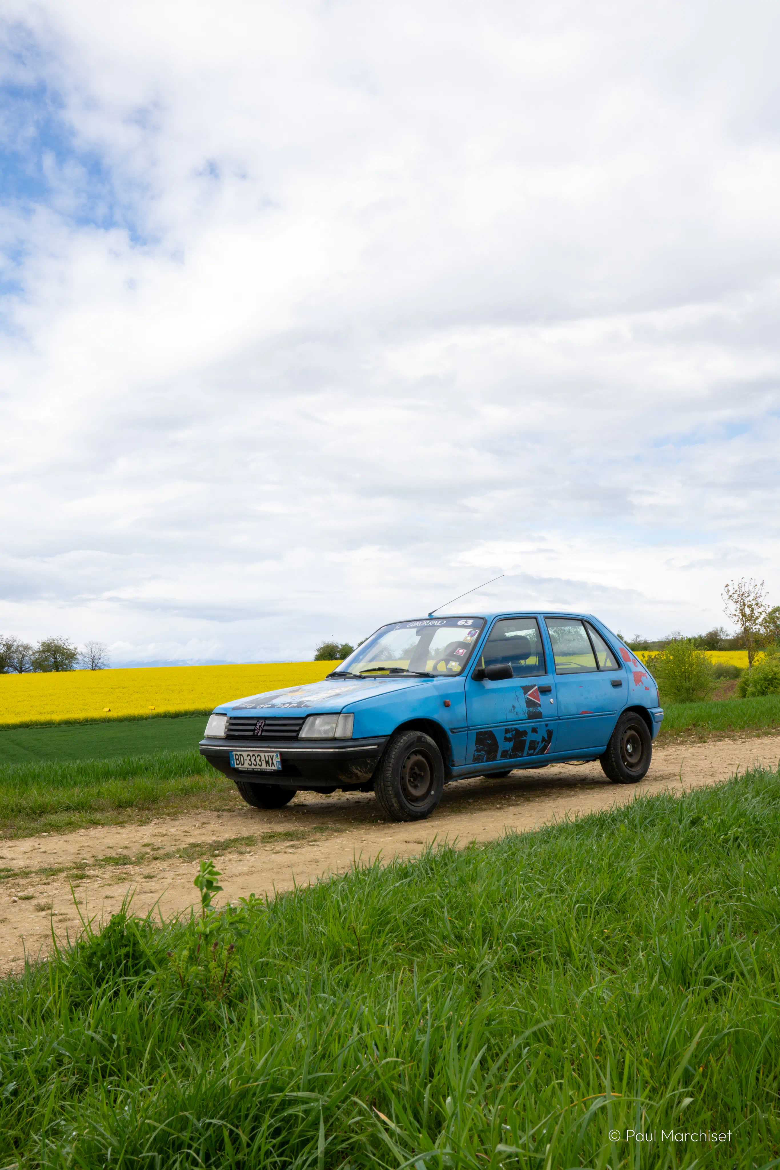 photo de notre peugeot 205 bleue devant un champs de colza jaune vif